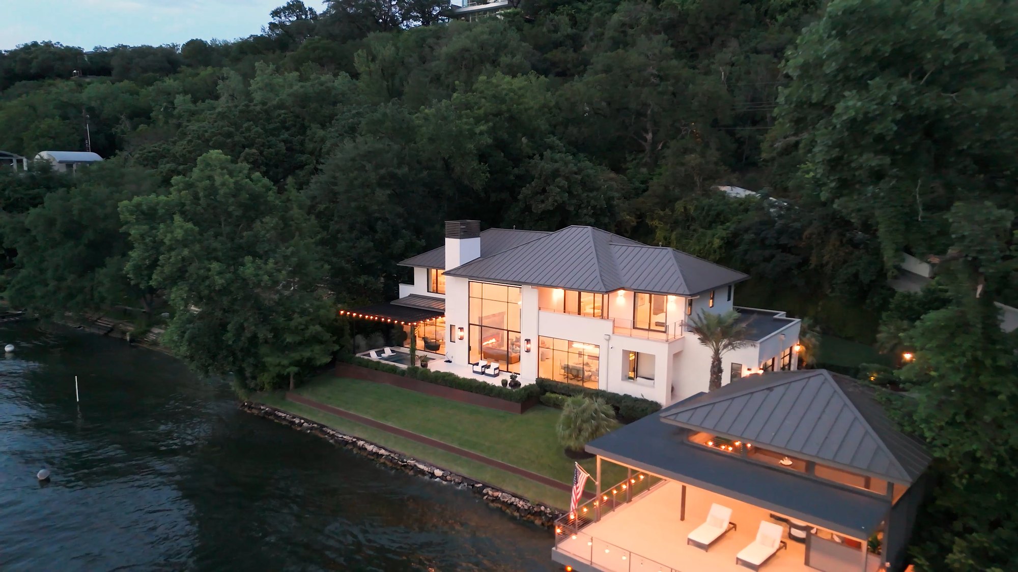 Aerial view of a modern glass-walled home on Lake Austin at dusk, its boathouse and terrace lit