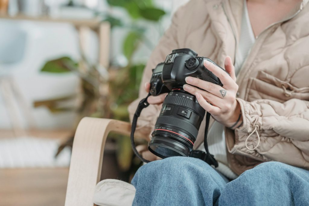 A person in casual attire holding a DSLR camera, sitting indoors